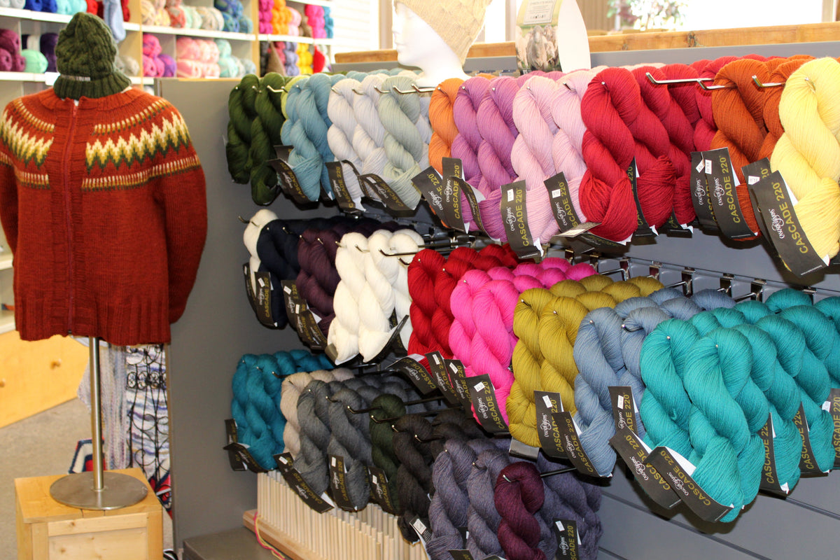 A retail display of colorful yarn skeins on wall racks, with a red knit sweater displayed on a mannequin wearing a green knit hat. The background shows shelves filled with more yarn in a bright store setting.